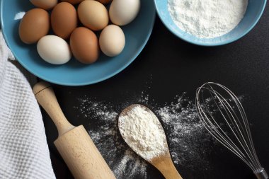 Hamurun hazırlanması. Un, yumurta, çırpma, haddeleme ve siyah masaüzerinde bir mutfak havlusu. Tahta bir kaşıkta bir demet un, mavi tabakta bir kase un ve yumurta. Pişirme maddesi üst görünümü