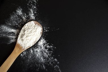 Tahta kaşıkta beyaz buğday unu yığını. Un siyah masaya serpilir. Koyu bir arka plan üzerinde un. Pişirme arka plan. Hamurun hazırlanması. Üst te. Lezzetli fotoğraf. Horizotal görüntü