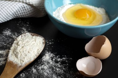 Un, yumurta ve mutfak havlusu. Koyu arka plan üzerinde hamur hazırlanması. Tahta bir kaşıkta bir demet un, bir kase un ve yumurta kabuğunda kırık bir yumurta. Siyah masaya serpişne un