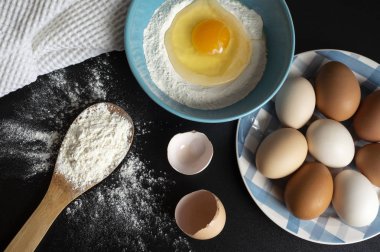 Un, yumurta ve mutfak havlusu. Koyu arka plan üzerinde hamur hazırlanması. Tahta bir kaşıkta bir demet un, bir kase un ve yumurta kabuğunda kırık bir yumurta. Un siyah masaya serpilir. Sayfanın Üstü