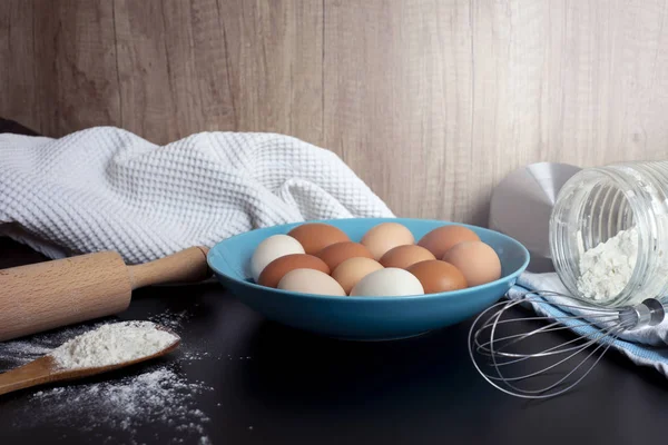 Preparation of the dough. Flour, eggs, whisk, rolling pin and a kitchen towel on the black table. A bunch of flour on a wooden spoon and lots of eggs on the blue plate. Baking reciepe and ingredients.