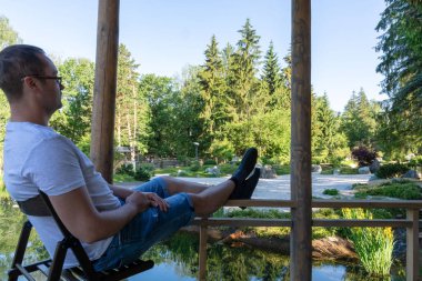 Genç bir adam Şafağı karşılar, ahşap katlanır sandalyede oturan bacaklarını ileriye doğru uzatarak ve onları Korkulmaya koyarak