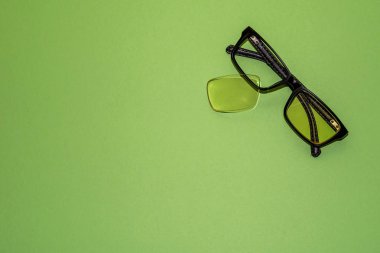 Kırık camlar yeşil bir yüzeyde yatıyor. Yakındaki çerçeveden düşmüş sarı bir lens yatıyor. Yukarıdan görüntüle