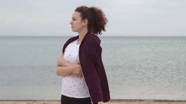 風が吹いている間に浜辺に立っている若い女性 若い女性は寒い天候でスウェットシャツで自分自身をドレスアップ — ストック動画