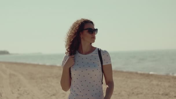 美丽的年轻女子卷曲的头发站在沙滩上 在阳光明媚的日子太阳镜 — 图库视频影像