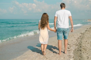 Güneşli bir günde el ele tutuşarak, sahilde yürüyen genç ve çekici bir çiftin arka görünümü