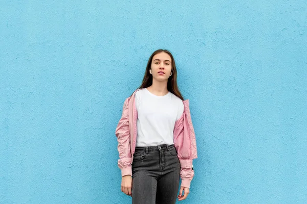 Beyaz tişörtlü, pastel pembe bombacı ceketli, mavi şehir duvarına dayanmış beyaz bir kızın portresi. Monokrom, moda minimal, şehir tarzı, model konsepti