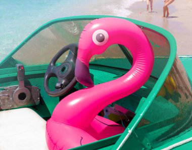 Şişirilebilir pembe flamingo, beyaz koltukta yeşil sürat teknesinde oturuyor. Güneşli bir yaz gününde, arka planda kumsalda koşan bacakları var. Aile tatili, tatiller, eğlence, eğlence