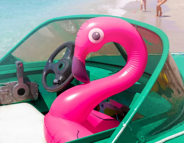 Şişirilebilir pembe flamingo, beyaz koltukta yeşil sürat teknesinde oturuyor. Güneşli bir yaz gününde, arka planda kumsalda koşan bacakları var. Aile tatili, tatiller, eğlence, eğlence