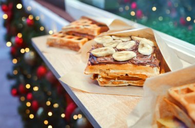 Noel pazarındaki sokak yemekhanesinde muz ve çikolata soslu Belçika waffle 'ları ve arka planda yeni yıl ışıkları var.