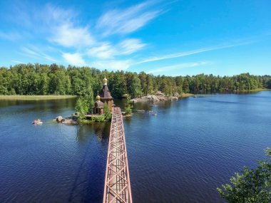 Nehir kıyıları boyunca bir orman ve açık bir yaz gününde ada üzerinde köprüsü olan bir kilise. Vuoksa 'daki St. Andrew Kilisesi. Rusya