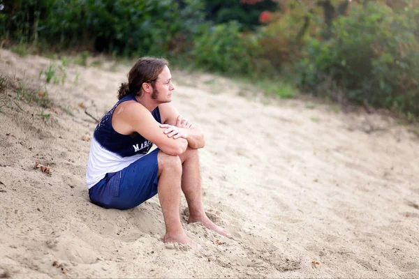 歳長い髪裸足の男性は 砂浜に座って水を見渡して — ストック写真