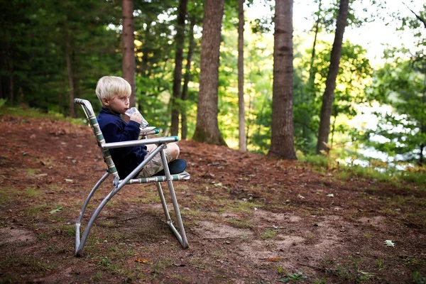 彼は湖の近くでキャンプしながらヴィンテージ芝生の椅子に座っているようにかわいい小さな金髪の子供がジュースを飲んで — ストック写真