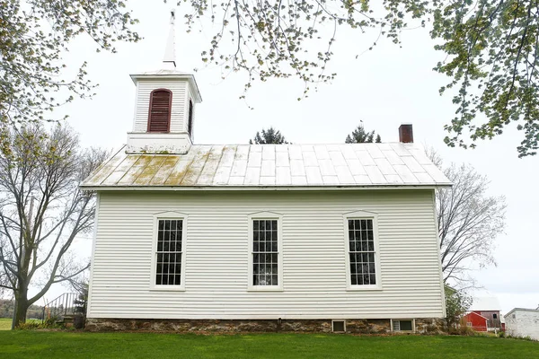 The white siding, stone foundation, and vertical grilled windows of a historic little white church in small town America, is framed by the leaves of tall shade trees.