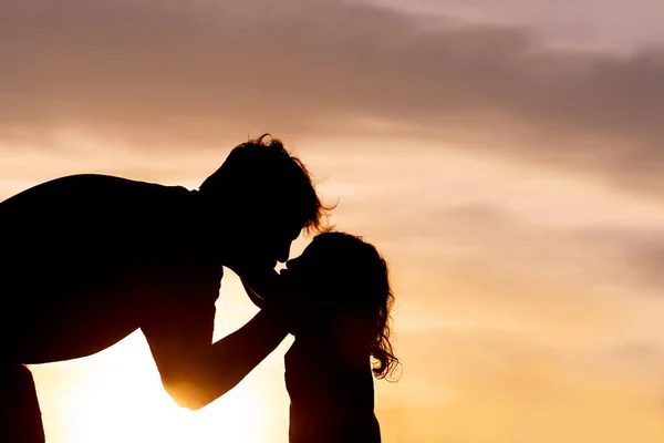 A silhouette of a loving father kissing and hugging his young toddler child at Sunset on a summer day.