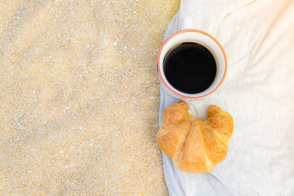 Black coffee and croissant on sand background, breakfast on the beach, food and drink concept.