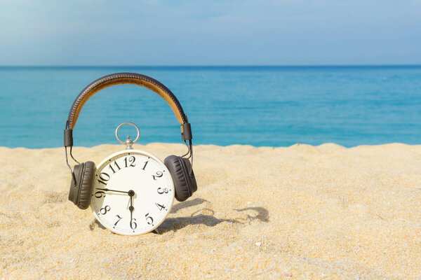 White alarm clock wearing brown headphones placed on the sand. Blue sea and sky as a background. Concept for relaxation, travel vacation and holidays with copy space. Summer time.