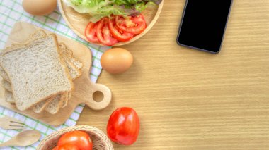 Ev yapımı sandviç kahvaltı hazırlanıyor. Tüm buğday ekmeği beyaz bir kumaşın üzerine yerleştirilen ahşap bir kesme tahtasında yığılmış, yeşil damalı desen. Ahşap masada akıllı telefon, dilim domates ve marul.