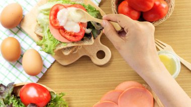 Ev yapımı sandviç kahvaltı hazırlanıyor. Kepekli ekmek beyaz bir kumaş, yeşil damalı desen üzerine yerleştirilen ahşap bir kesme tahtası üzerine yığılmış. Dilim domates ve marul ahşap bir plaka üzerine yerleştirilir. Taze domates, jambon, yumurta, kaşık ve çatal bir w yatıyordu