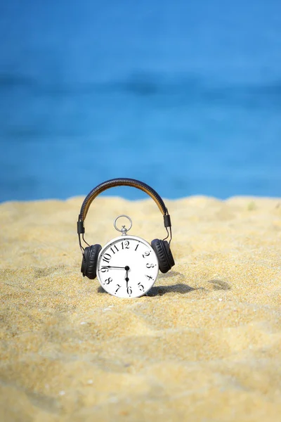 White alarm clock with brown headphones placed on the sand. Beautiful blue sea and sky as a background. Concept for relaxation, travel vacation and holidays with copy space. Summer time.