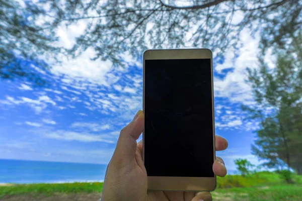 Close up young woman hand holding a modern black smartphone mock up in horizontal position with blank screen against beautiful blurred sea and sky background.