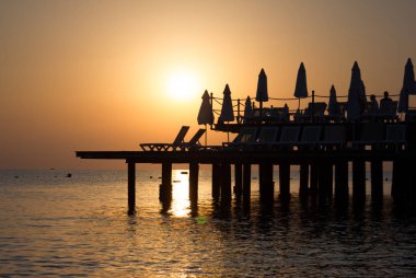 Sıcak turuncu ve altın tonları ile denizin güzel günbatımı manzaralı arka plan. Şezlong ve şemsiye iskelede vardır.
