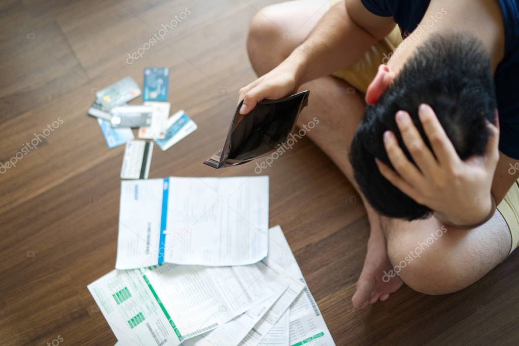 Top view of stressed young Asian man holding empty wallet and thinking about  finding money to pay credit card debt and all bills. He is holding head by another hand. Financial problem concept.