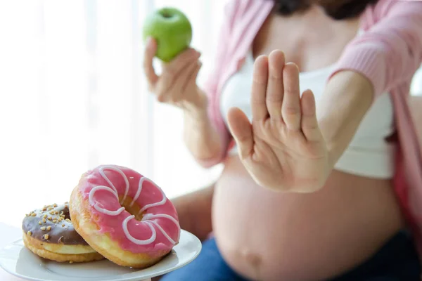 节食怀孕的女性拒绝自己最喜欢的垃圾食品 比如甜甜圈 而是选择绿色苹果 转而为自己的新生儿未来的健康状况 快乐的母亲和保健理念 — 图库照片