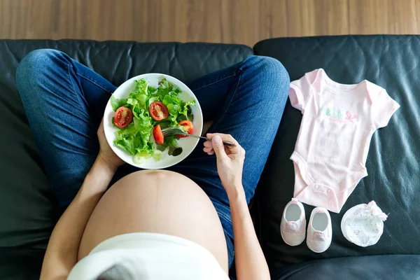 坐在沙发上的孕妇手里拿着沙拉碗 她未来出生的婴儿礼服放在她旁边的沙发上 在最上面的视野中看到 专注于沙拉 健康为母亲的概念 — 图库照片