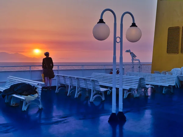 Young woman on ship looking at the sunset, blue deck white benches, yellow chimney, distant island mountains, Aegean sea, Greece.