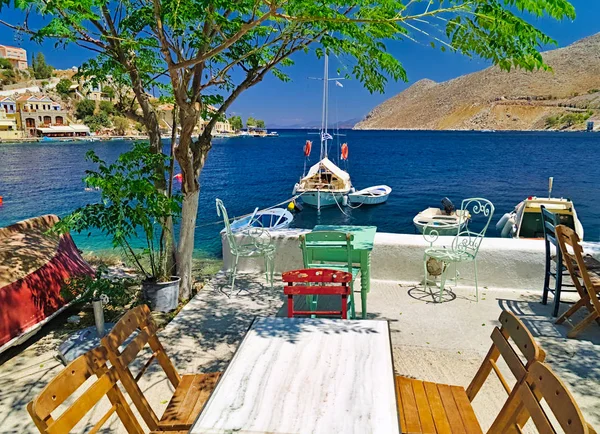 Symi ada limanı, masa ve sandalyeler, ağaç, deniz ve yelkenli tekne ile kafeden görünümü.