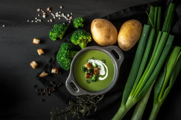 Spring detox broccoli green cream soup with potatoes and vegan cream in bowl on dark wooden board over black background, top view. Clean eating, dieting, vegan, vegetarian, healthy food concept