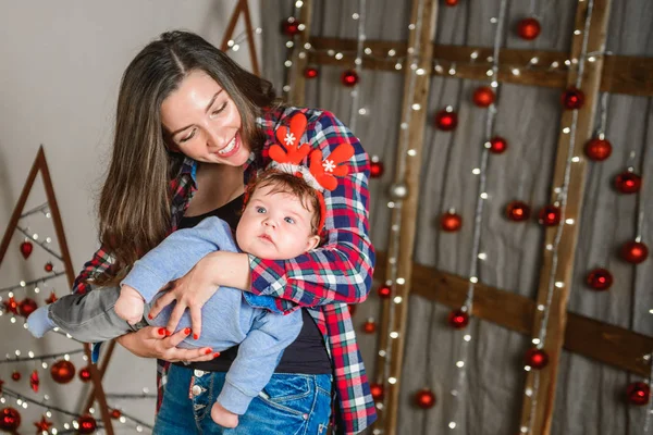 Mom and baby hug at the Christmas tree. Happy baby and his mom are looking at the frame. Mom with son hugging for Christmas. and have fun. newborn baby for the new year.