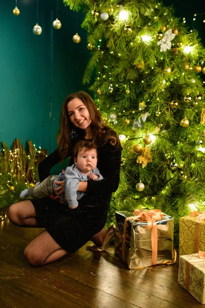 Mom and baby hug at the Christmas tree. Happy baby and his mom are looking at the frame. Mom with son hugging for Christmas. and have fun. newborn baby for the new year.