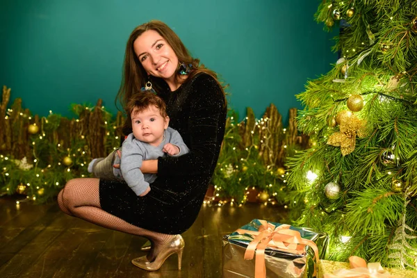 Mom and baby hug at the Christmas tree. Happy baby and his mom are looking at the frame. Mom with son hugging for Christmas. and have fun. newborn baby for the new year.