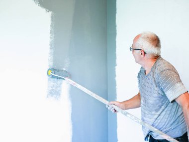 Yaşlı bir adam silindirle bir duvar boyuyor. Kıdemli adam tamirat yapar. Duvarları silindirle boyamak. Eski kıyafet. Beyaz bir duvarda gri bir rüzgar. ev onarımı