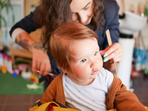 První sestřih. Roztomilé blondýny batole je šťastný, že na účes s profesionálním baby hairdresser. Blonďatý chlapeček se stříhá v kadeřnictví. Ruce kadeřníka dělat účes f — Stock fotografie