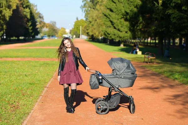 Anneyi bebek arabasıyla gezintiye çıkarmış. Parkta bebekle yürüyen genç bir anne. Güzel bir gülümseme ve keyifli bir ruh hali, hayattan memnun.. — Stok fotoğraf