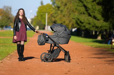 Bebek arabalı genç bir anne. Aile, çocuk ve ebeveynlik kavramı - mutlu anne parkta bir bebek arabasıyla yürüyor.