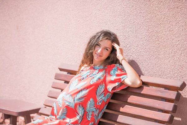 Het zwangere meisje koestert zich in de zon. Het concept van warme vrije tijd - een mooi zwanger meisje met lang haar, koesterend in een rode jurk. zittend op een bank. roze achtergrond. plaats van schrijven — Stockfoto