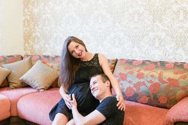 Glückliches Paar erwartet ein Baby. glücklicher Lebensstil der Familie. junges Paar erwartet ein Baby. Schwangere liegt auf Sofa und blickt mit ihrem Mann in die Kamera, umarmt sich und lächelt glücklich. — Stockfoto