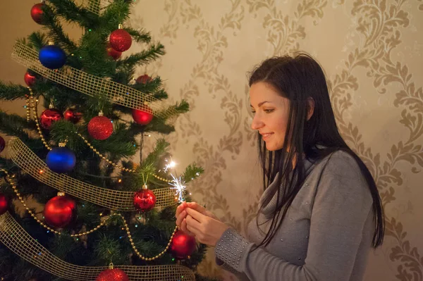 Egy lány egy csillagszóróval a karácsonyfa mellett. Egy vidám fiatal nő, aranyos mosollyal, kötött pulóverben áll és egy csillogó csillagszórót tart a kezében a Chri hátterében. — Stock Fotó