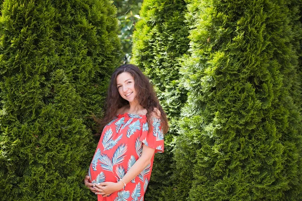 Těhotná dívka v blízkosti krásné zeleně. Krásná těhotná žena v šatech a dotýkající se břicha stojí v blízkosti thuja. Budoucí matka v módních šatech. Těhotné břicho s rukama. — Stock fotografie