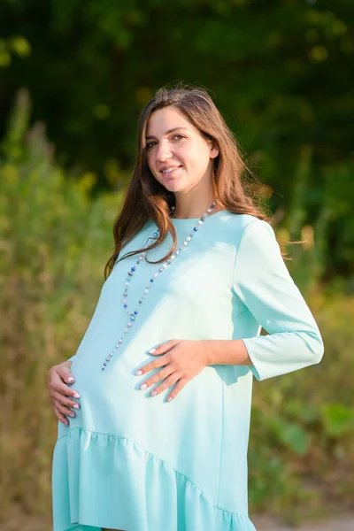 Terhes lány türkiz ruhában. Terhes nő napközis ruhában sétálgat a parkban. A lány mosolyog. kényelmes ruhák terhes nők számára. — Stock Fotó