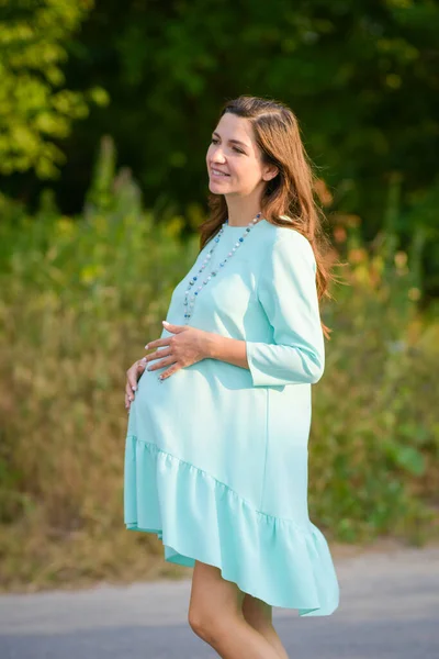 청록색 드레스를 입은 임산부입니다. 낮에 임신 한 여자 가 공원에서 놀고 있어요. 소녀는 미소를 짓습니다. 임신부들을 위한 편안 한 옷. — 스톡 사진