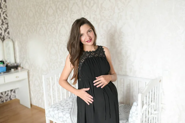 Pregnant girl near the bed for a child. kravatka white color for the child. preparing for the birth of a child. photo of a pregnant woman standing near the bed and hugging her belly — Stock Photo, Image