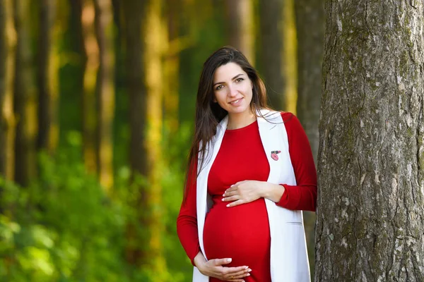 Ciąża w czerwieni. Portret pięknej kobiety w ciąży w czerwonej sukience w kwitnącym wiosennym parku — Zdjęcie stockowe