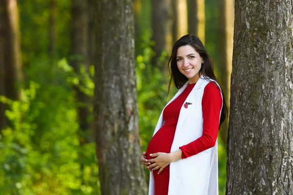 빨간 드레스를 입은 임신 한 여자. 꽃 이 만 발 한 봄 공원에서 붉은 옷을 입은 아름다운 임신부 의사 진 — 스톡 사진