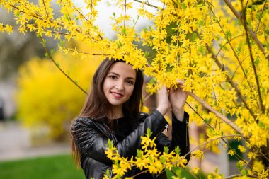 Sonbahar bahçesinde esmer poz veriyor. Genç ve güzel esmer kız gülümsüyor. Açık havada. Yeşil yapraklı ağaçlar. Bahçe, park. Güneşli bir gün. sarı yapraklar