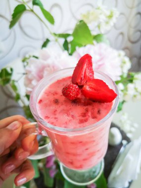 Bir kızın elinde çilekli smoothie. Böğürtlenli pembe kokteyl içen bir kadın, çiçeklerin arka planına karşı bir kokteyl, dikey fotoğraf..